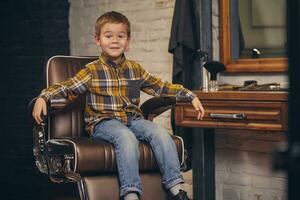 retrato do uma à moda pequeno Garoto vestido dentro camisa e jeans dentro a barbearia, sentado dentro uma cadeira contra a barbearia local de trabalho foto