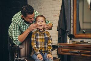 à moda pequeno criança sentado em cadeira às barbearia com dele jovem pai em fundo foto