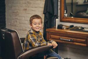 retrato do uma à moda pequeno Garoto vestido dentro camisa e jeans dentro a barbearia, sentado dentro uma cadeira contra a barbearia local de trabalho foto