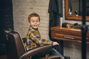 retrato do uma à moda pequeno Garoto vestido dentro camisa e jeans dentro a barbearia, sentado dentro uma cadeira contra a barbearia local de trabalho foto