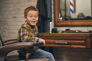 retrato do uma à moda pequeno Garoto vestido dentro camisa e jeans dentro a barbearia, sentado dentro uma cadeira contra a barbearia local de trabalho foto