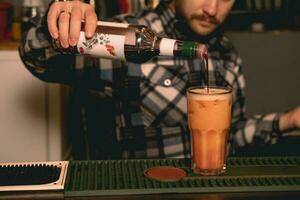 barman preparando tequila nascer do sol coquetel, adicionando romã xarope para dentro vidro com beber foto