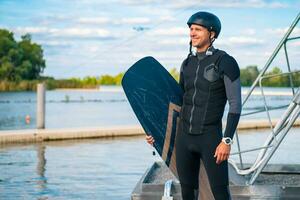 sorridente praticante de wakeboard dentro roupa de mergulho e capacete com borda em pé em cais foto