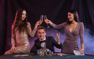 homem jogando pôquer às cassino sentado às mesa com pilhas do salgadinhos, dinheiro, cartões. a comemorar ganhar com dois mulheres. preto, fumaça fundo. fechar-se. foto