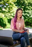 mulher escrevendo dentro uma caderno sentado em uma de madeira Banco dentro a parque. menina trabalhando ao ar livre em portátil computador, cópia de espaço. foto