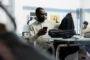 africano americano homem escolhendo casual Jaqueta dentro roupas loja e verificação preço em local na rede Internet usando Móvel telefone. departamento Shopping cliente navegando vestuário prateleira e examinando boutique aplicativo foto