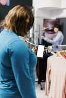 africano americano cliente segurando Smartphone com branco tela dentro compras shopping center, verificação moda loja aplicativo. à moda mulher comprando elegante mercadoria dentro moderno boutique. tecnologia conceito foto