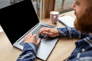 cortada imagem do jovem homem conversando através da netbook durante trabalhos pausa dentro café comprar, masculino sentado dentro frente aberto computador portátil computador com em branco cópia de espaço tela. foto