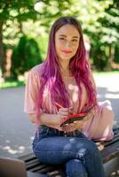 mulher escrevendo dentro uma caderno sentado em uma de madeira Banco dentro a parque. menina trabalhando ao ar livre em portátil computador, cópia de espaço. foto