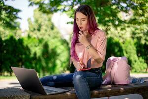 mulher escrevendo dentro uma caderno sentado em uma de madeira Banco dentro a parque. menina trabalhando ao ar livre em portátil computador, cópia de espaço. foto