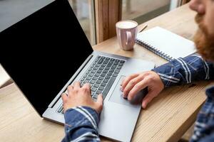 cortada imagem do jovem homem conversando através da netbook durante trabalhos pausa dentro café comprar, masculino sentado dentro frente aberto computador portátil computador com em branco cópia de espaço tela. foto