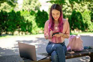 mulher escrevendo dentro uma caderno sentado em uma de madeira Banco dentro a parque. menina trabalhando ao ar livre em portátil computador, cópia de espaço. foto