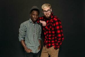 estúdio tiro do dois à moda jovem homens tendo Diversão. bonito barbudo hipster dentro uma camisa dentro uma cela em pé Próximo para dele afro-americano amigo dentro chapéu contra uma Sombrio fundo. foto