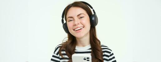 retrato do lindo jovem mulher escuta para música dentro sem fio fones de ouvido, usa dela Móvel telefone, sorrisos e parece feliz, isolado em branco fundo foto