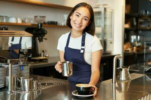 lindo jovem fêmea barista, fazer capuccino, derramando cozido no vapor leite para café com leite arte para dentro xícara, dar seu ordem atrás contador foto