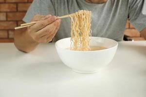 homem tailandês casual com fome usa os pauzinhos para comer macarrão instantâneo quente no copo branco durante as pausas para o almoço, rápido, saboroso e barato. refeição tradicional asiática saudável fast-food do estilo de vida japonês e chinês. foto