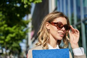 fechar acima retrato do sorridente jovem mulher dentro oculos de sol, corporativo terno, segurando uma pasta com trabalhos documentos, indo para a entrevista, em pé ao ar livre foto