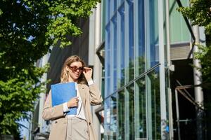retrato do confiante corporativo mulher indo para escritório com uma pasta, caminhando em rua em ensolarado dia dentro terno e oculos de sol foto