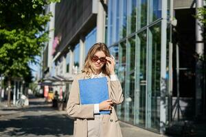 retrato do corporativo mulher dentro oculos de sol e bege terno, segurando azul pasta com escritório documentos, indo para trabalhar, caminhando em rua em ensolarado dia foto