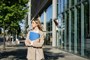 trabalhos e corporativo pessoas. confiante empresária segurando uma azul pasta com o negócio documentos, caminhando em rua foto