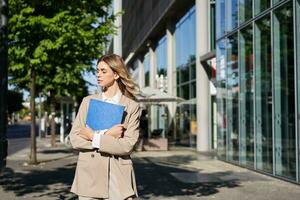 trabalhos e corporativo pessoas. confiante empresária segurando uma azul pasta com o negócio documentos, caminhando em rua foto