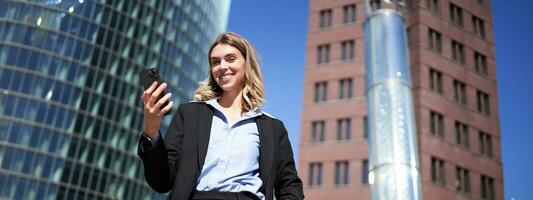 baixo ângulo tiro do empresária dentro terno, em pé em rua e olhando às Móvel telefone, segurando Smartphone foto