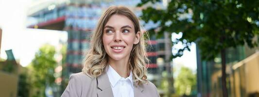 fechar acima retrato do sorridente, lindo mulher 25 anos velho, vestindo corporativo roupas, olhando feliz, em pé lado de fora em rua foto