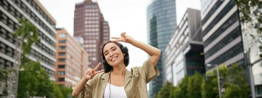 feliz ásia mulher dentro fones de ouvido, ouvindo música e dançando em rua do cidade Centro, sorridente com mãos acima foto