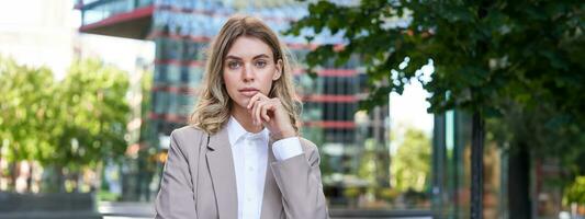 retrato do confiante corporativo mulher, jovem fêmea CEO Gerente Veja às Câmera, posando dentro bege terno perto escritório edifícios, em pé em rua foto