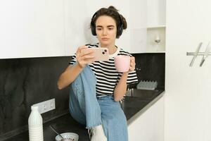 estilo de vida conceito. jovem moderno mulher relógios filme em Smartphone, vestindo fones de ouvido, bebidas café ou chá dentro a cozinha foto