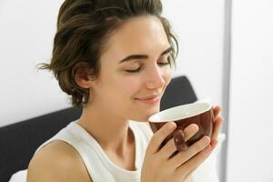 fechar acima retrato do morena, sorridente mulher, cheirando dela café dentro xícara, acordando acima e bebendo cappuccino dentro cama foto