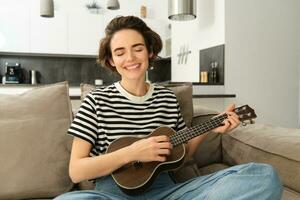jovem feliz mulher sentado em sofá e jogando cavaquinho, cantando e desfrutando Aprendendo Novo musical instrumento foto