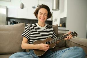 apaixonado jovem mulher, músico jogando ukulele às lar, cantando com alegria, sentado em sofá dentro vivo quarto foto