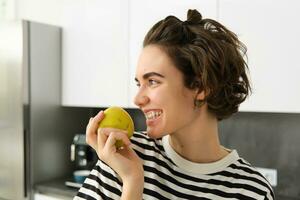 fechar acima retrato do fofa jovem moderno mulher, comendo verde maçã e olhando aparte, sorridente enquanto morder uma fruta, tendo uma lanche dentro a cozinha foto