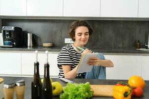 sorridente morena mulher dentro cozinha, escrevendo baixa refeição Lista para semana, lendo receita, cozinhando salada, vegetal refeição às casa foto