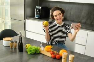 sorridente jovem mulher, Comida blogueiro gravação uma vídeo em Smartphone com comida, cozinhando e fazer selfie, levando As fotos durante refeição preparação, sentado perto legumes e segurando a maçã