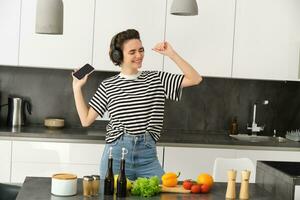 retrato do feliz dançando mulher, cozinhando Comida dentro a cozinha, ouvindo música dentro sem fio fones de ouvido, cortar legumes para salada, preparando saudável Comida e desfrutando favorito música foto