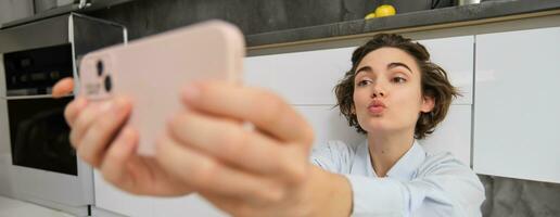 fofa morena mulher leva selfie em Smartphone, franzidos lábios e poses para foto em Móvel telefone, senta em cozinha chão às casa