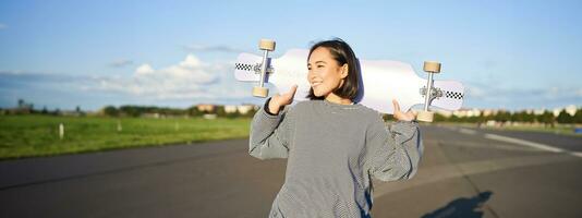 estilo de vida e pessoas. jovem ásia menina posando com Longboard, patinação em dela cruzador. sorridente mulher segurando skate em ombros foto