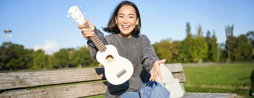 feliz ásia menina mostra cavaquinho, demonstra dela Novo musical instrumento, senta em Banco dentro parque e tocam música foto