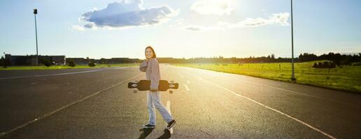 jovem skatista garota, adolescente patinação em cruzador, segurando Longboard e caminhando em concreto esvaziar estrada foto