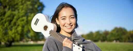 retrato do lindo sorridente menina com cavaquinho, ásia mulher com musical instrumento posando ao ar livre dentro verde parque foto