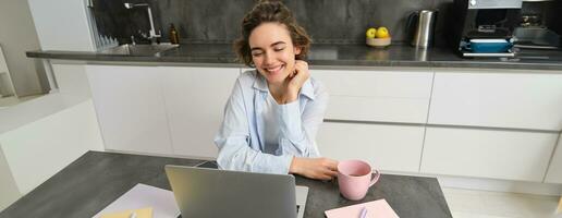 jovem mulher trabalhador autonomo trabalho a partir de lar, parece às computador portátil tela e sorrisos, faz dever de casa dentro cozinha foto