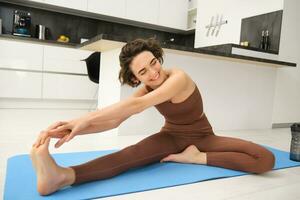 retrato do em forma morena mulher, ginástica instrutor fazendo exercícios às lar, alongar dela pernas, aquece antes ioga Treinamento dentro dela quarto foto