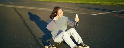 feliz ásia menina senta em skate, leva selfie com Longboard, faz fofa rostos, ensolarado dia ao ar livre foto