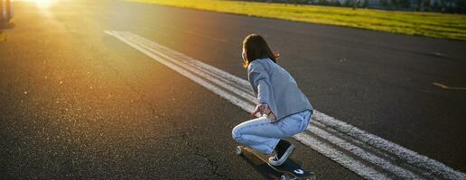 lado Visão do lindo ásia menina em skate, equitação dela cruzador para a Sol em a esvaziar estrada. feliz jovem skatista desfrutando ensolarado dia em dela patim foto