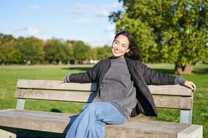 retrato do jovem mulher dentro ao ar livre roupas, sentado em Banco relaxado, sorridente e desfrutando Visão em verde parque foto