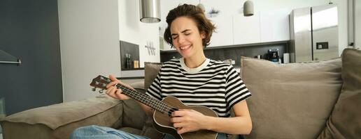 retrato do jovem moderno mulher, aluna jogando ukulele às lar, sentado com pequeno guitarra, cantando e sentindo-me feliz, sentado em sofá foto