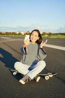 feliz adolescente ásia menina leva selfie, vídeo chats em Smartphone aplicativo enquanto senta em skate, skatista registros vlog enquanto senta em Longboard foto