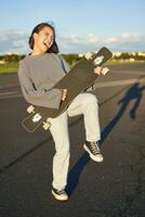engraçado ásia menina desfrutando patinação, segurando skate gostar guitarra e sombra jogando, tendo Diversão ao ar livre foto